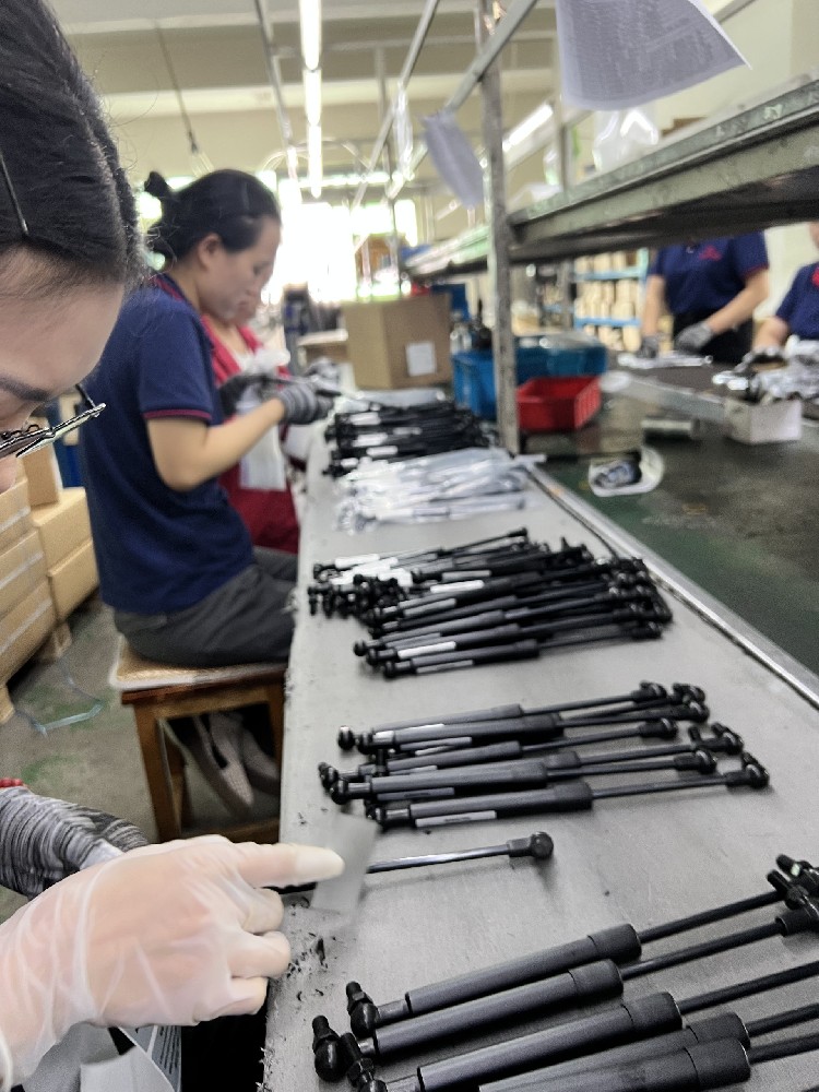Workers handling gas springs at a workbench with rows of black gas springs.
